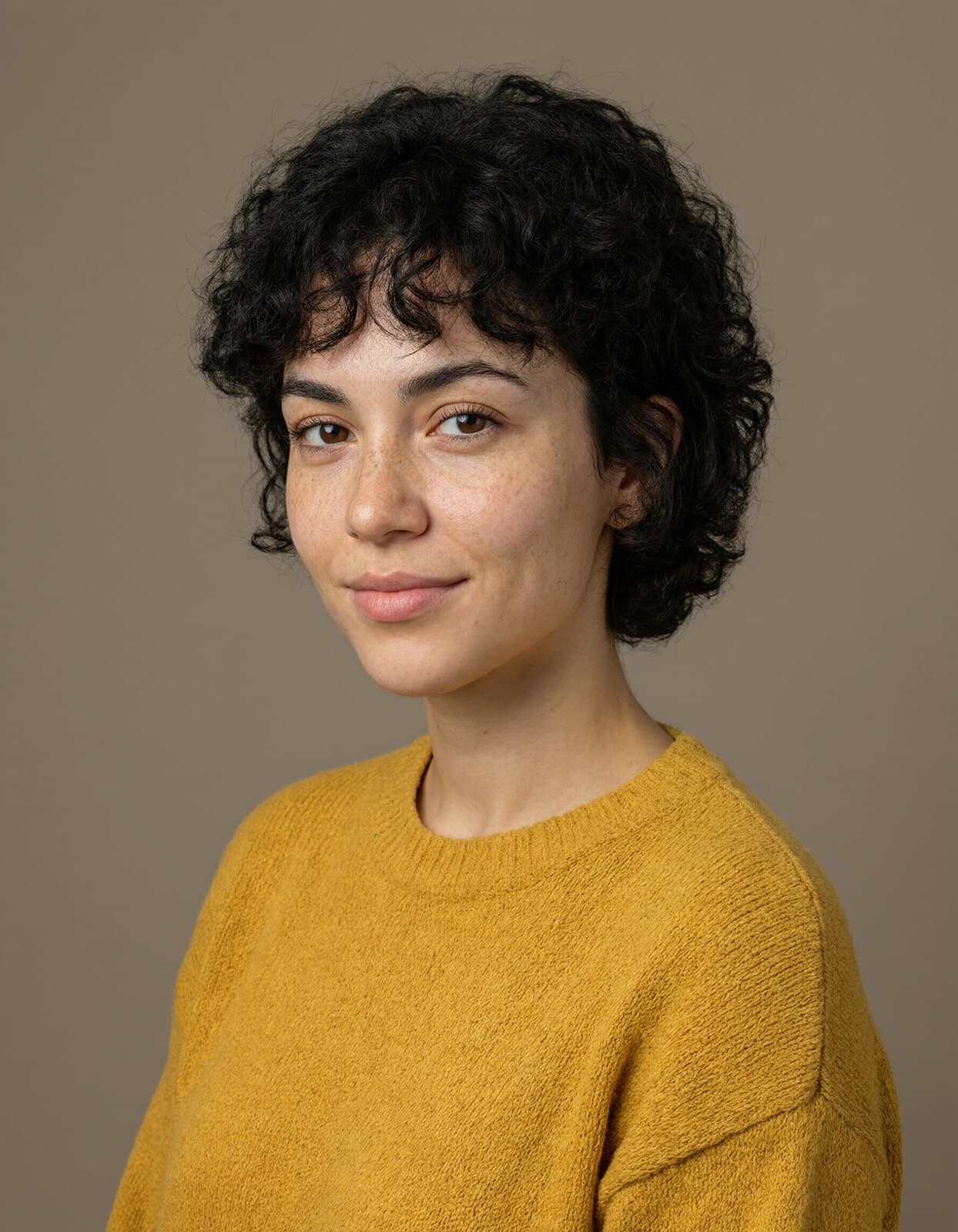 Ein Studioporträt einer Frau mit kurzen, lockigen schwarzen Haaren in einem senfgelben Pullover